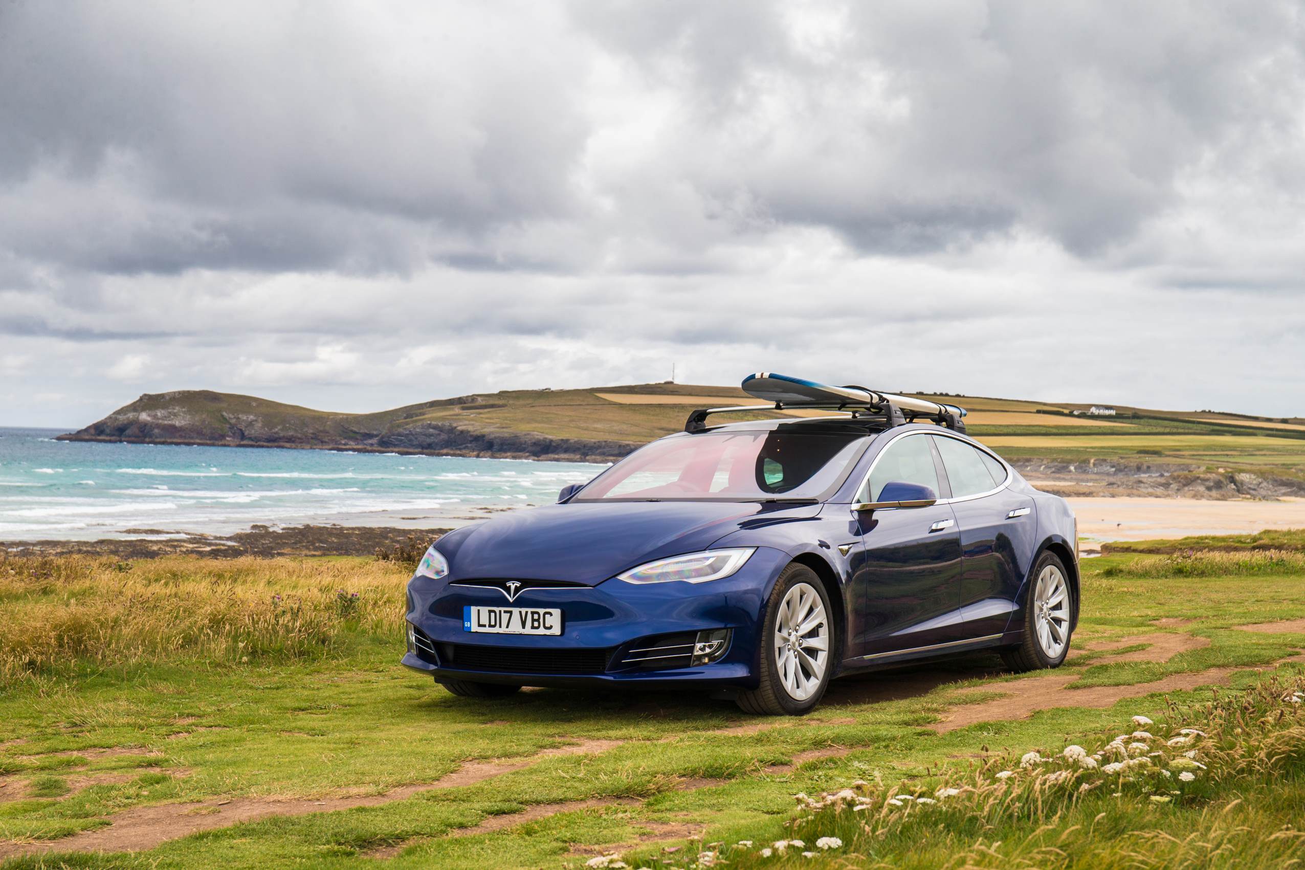 Blue Tesla Model S on grass by sea with surfboard on roof